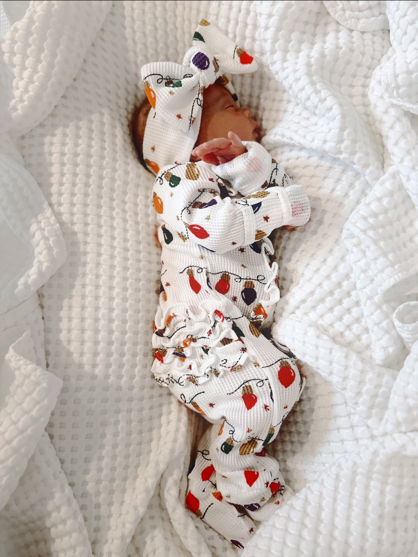 Newborn wearing festive patterned outfit and large bow, peacefully sleeping on white blanket.