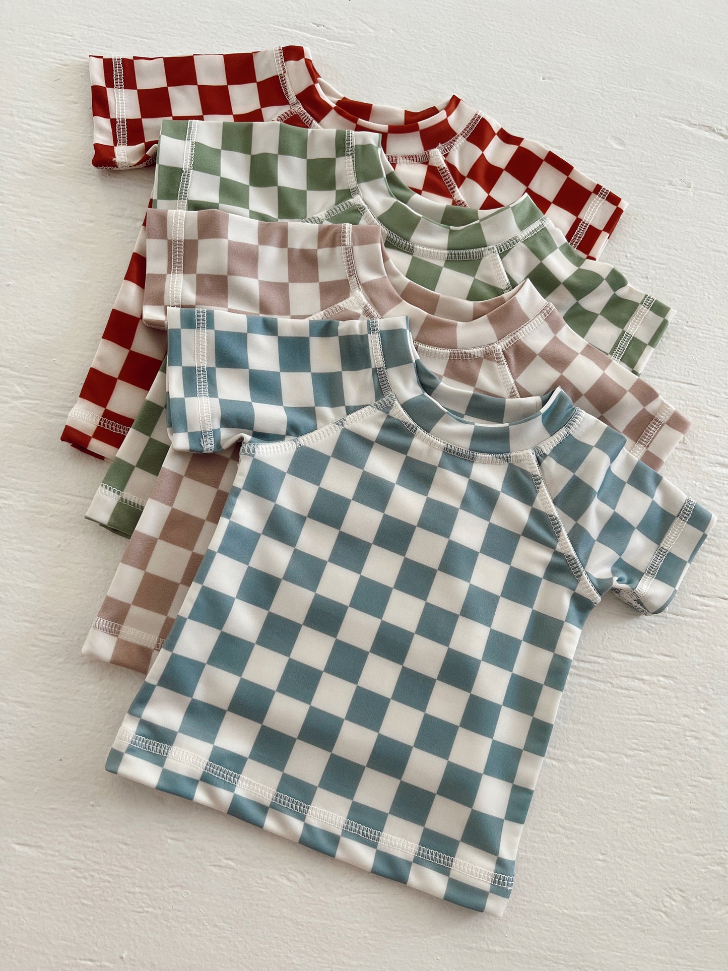 Four checkered t-shirts in red, green, brown, and blue stacked on a white surface.