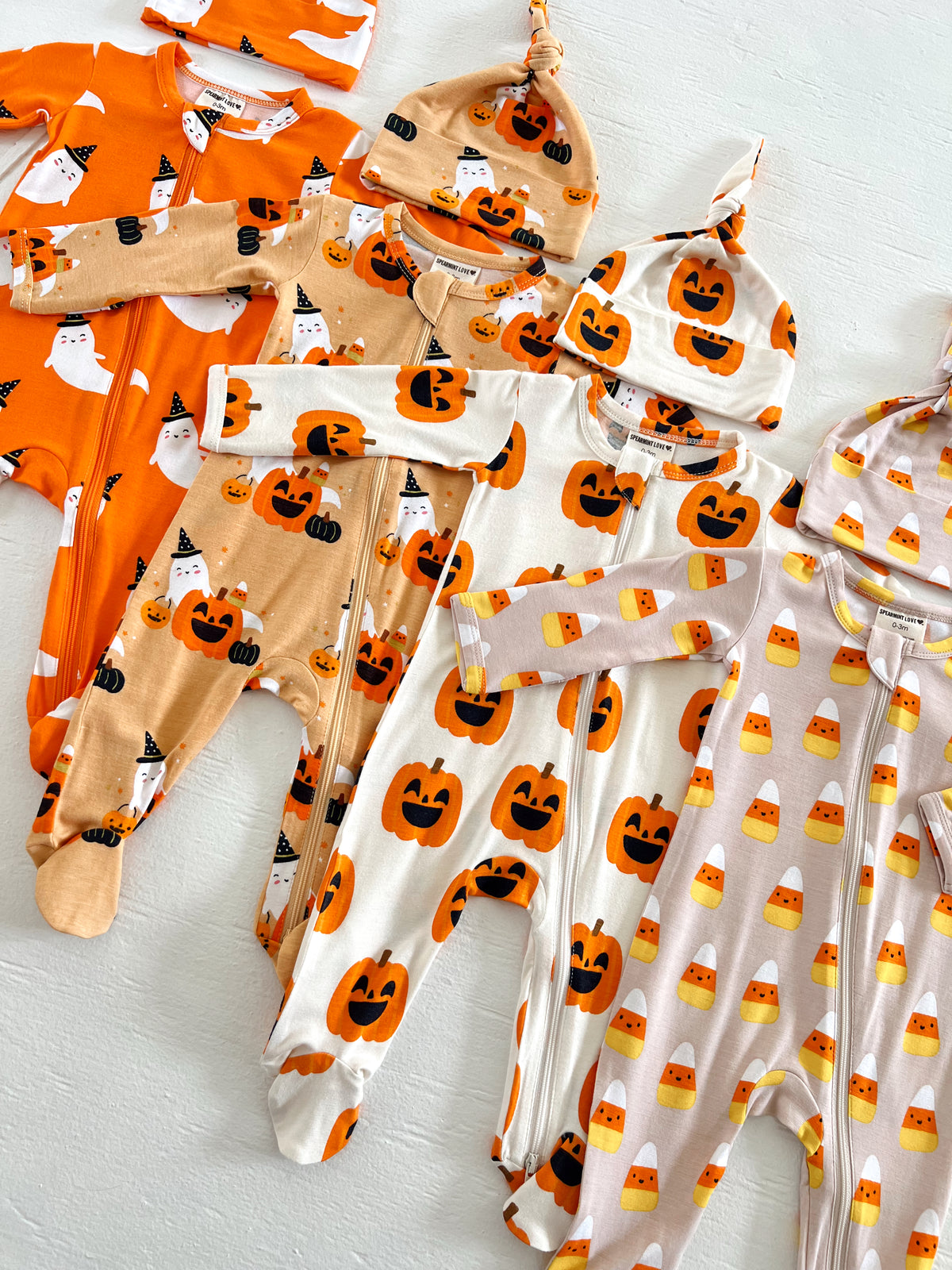 Colorful baby Halloween pajamas and hats with pumpkin, ghost, and candy corn patterns displayed on a light background.