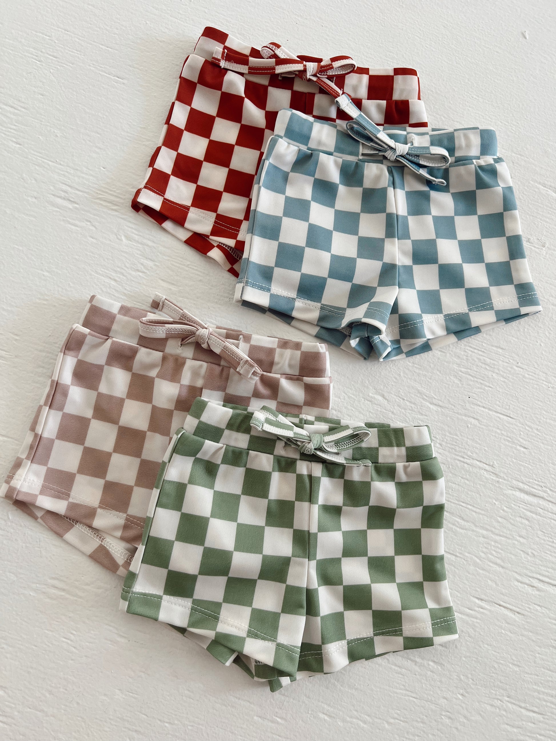 Four pairs of checked shorts in red, blue, green, and brown laid out on a white surface.