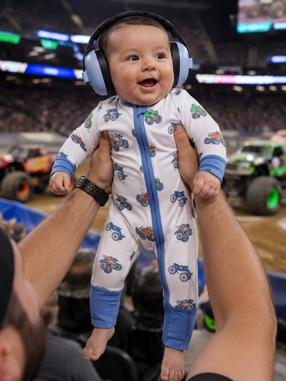 Baby in noise-canceling headphones smiles while being held at a monster truck event, wearing a playful, patterned onesie.
