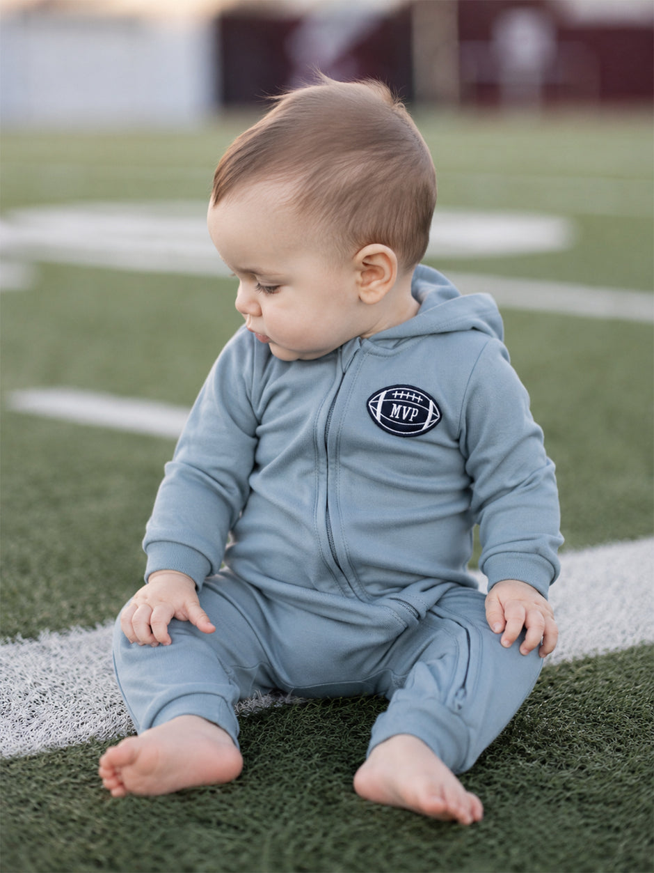 Baby wearing a light blue sports onesie sits on a football field, looking to the side.