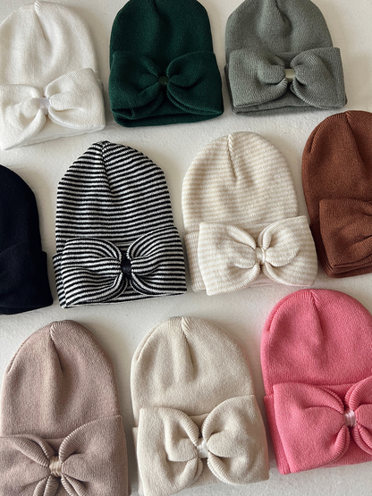 Assorted beanies in various colors and styles, featuring bows, arranged on a white background.