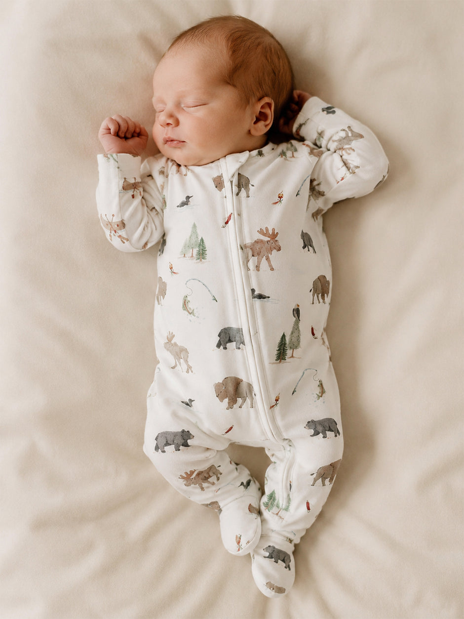 Sleeping baby in animal-patterned onesie on a soft blanket, surrounded by nature-themed illustrations.