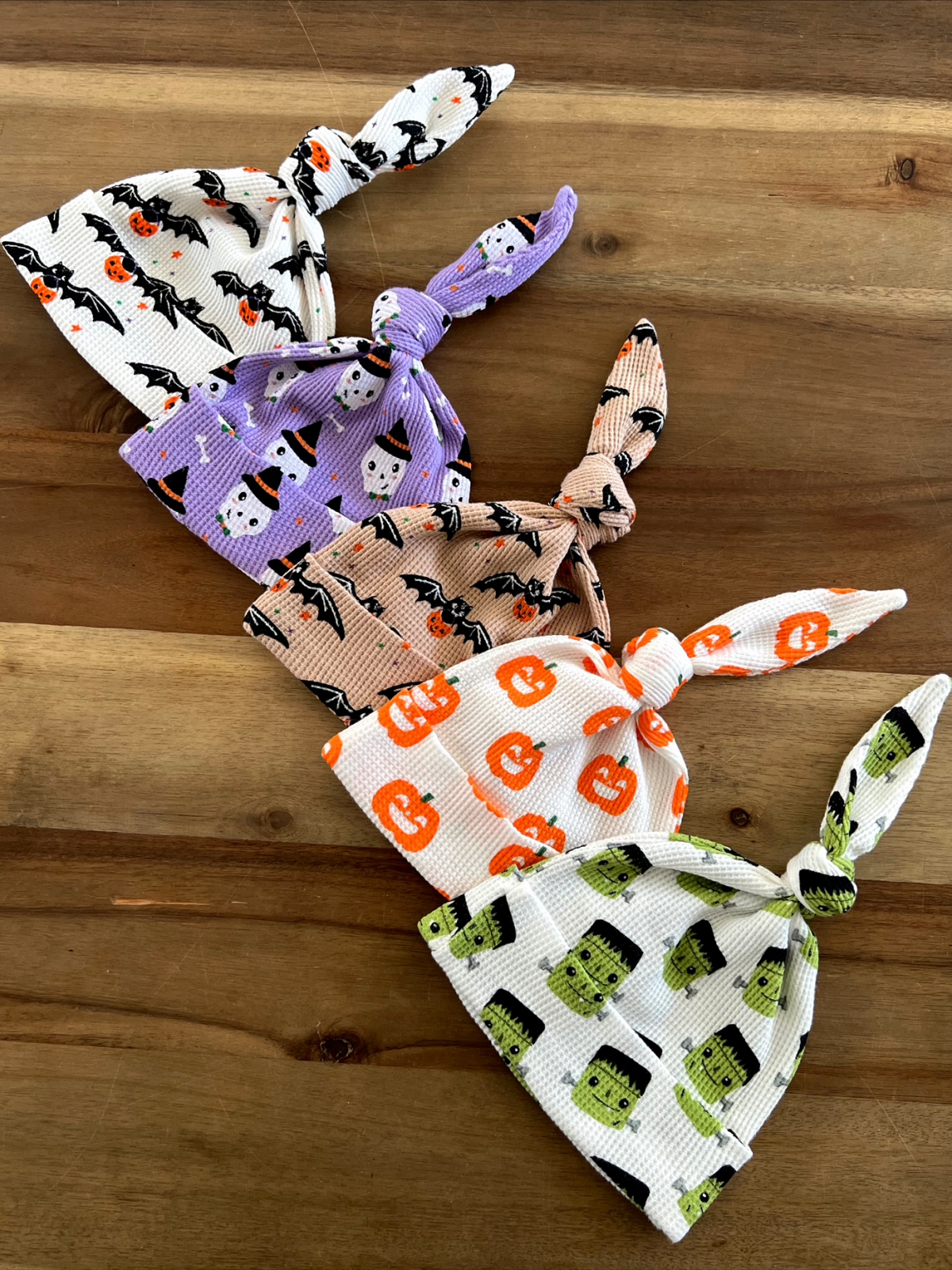 Five Halloween-themed knotted caps with patterns of bats, pumpkins, ghosts, and Frankenstein on a wooden surface.