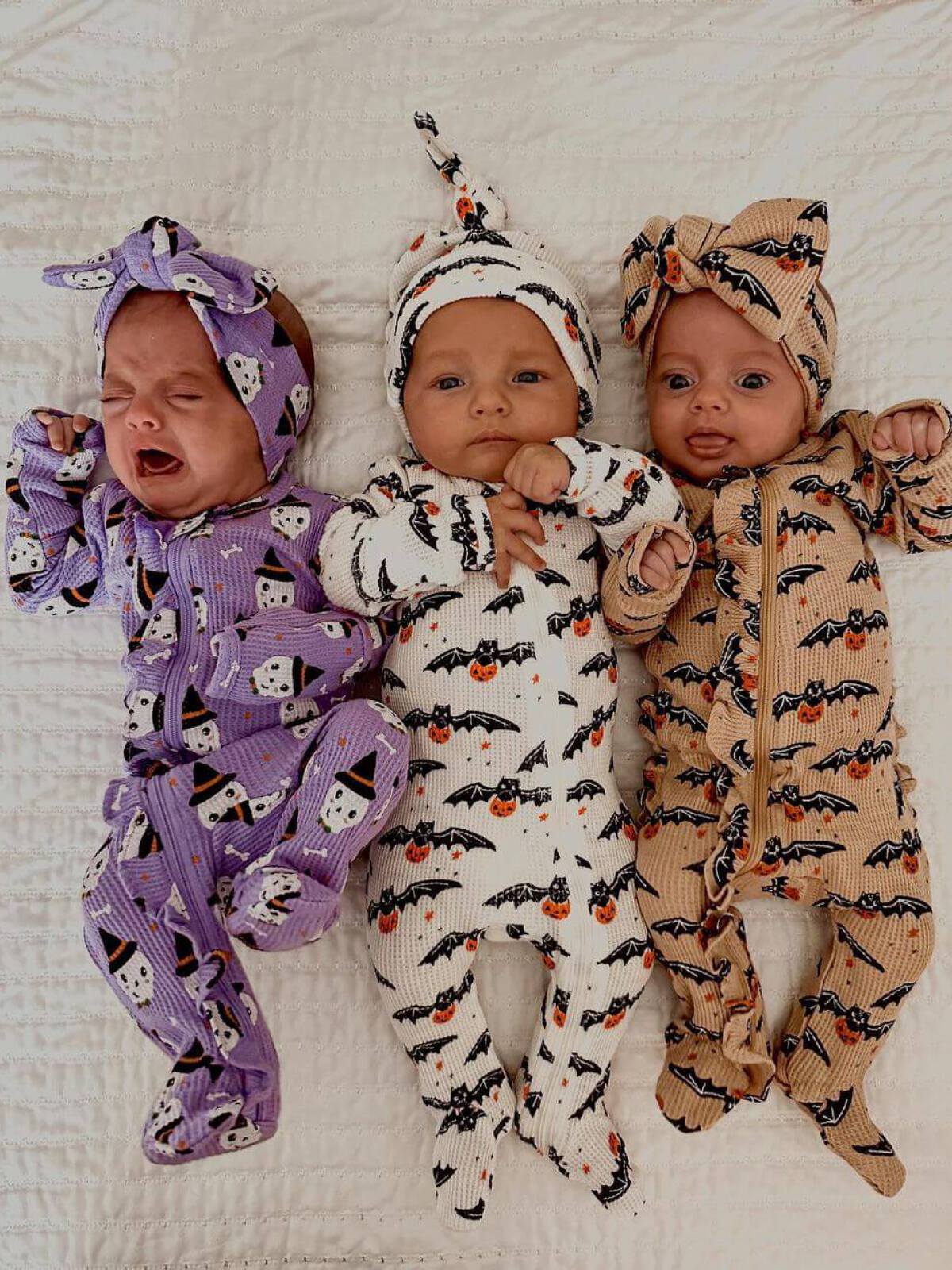 Three babies in Halloween-themed pajamas pose on a white blanket; one is crying, others are smiling.