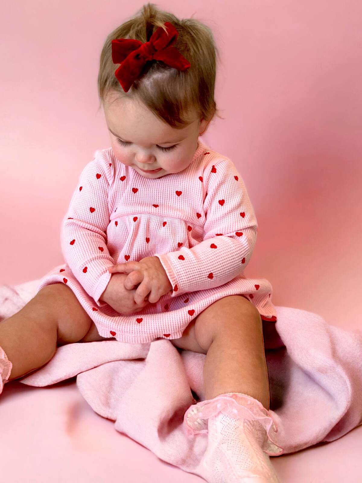 Baby girl wearing a heart-patterned dress and red bow, sitting on a pink blanket, focused on her hands.