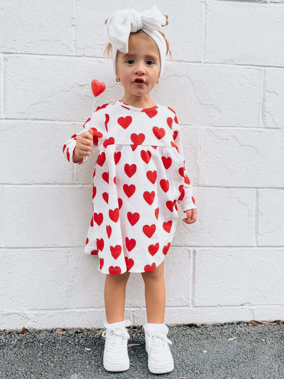 Smiling child in a heart-patterned dress, holding a heart-shaped lollipop against a white wall.