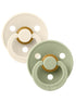 Two pacifiers, one in cream and one in sage green, both with round designs and a small handle.