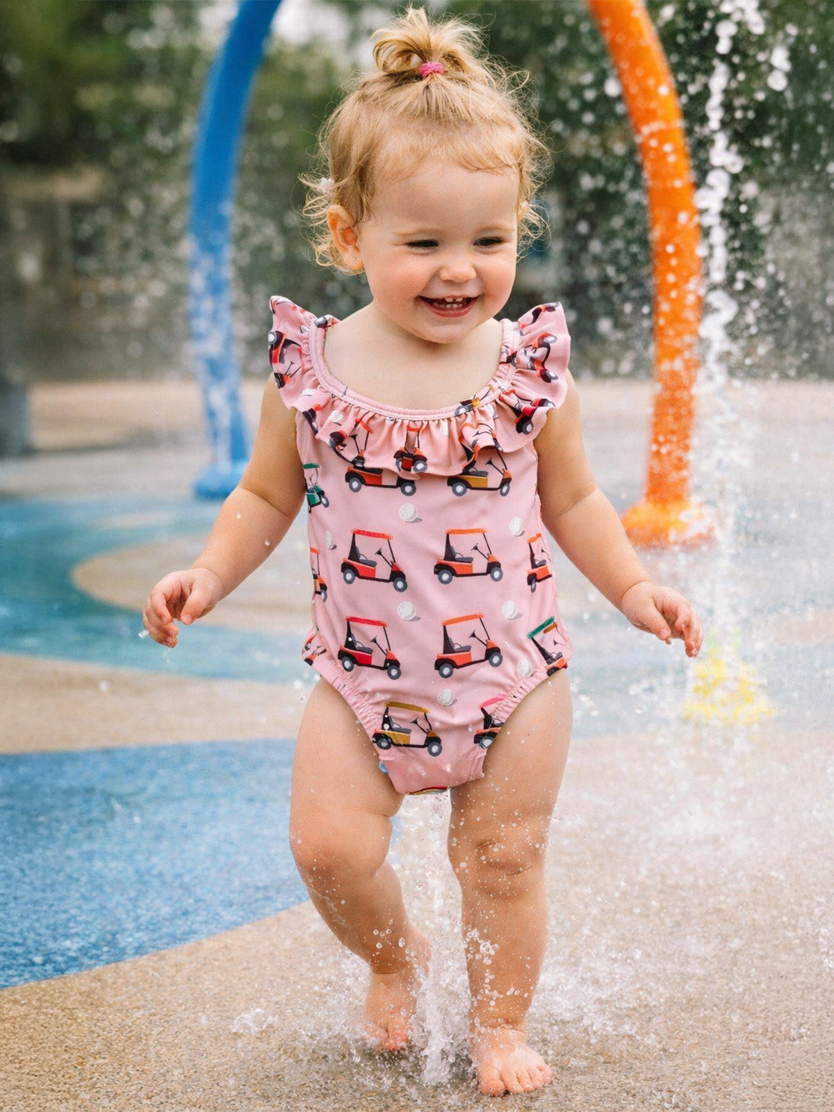 Smiling toddler in a pink swimsuit with ruffles, playing in a splash pad surrounded by colorful water features.