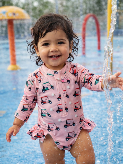 Smiling toddler in a pink swimsuit with golf cart prints playing in a splash pad. Water sprays around them.