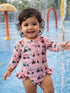 Smiling toddler in a pink swimsuit with golf cart prints playing in a splash pad. Water sprays around them.