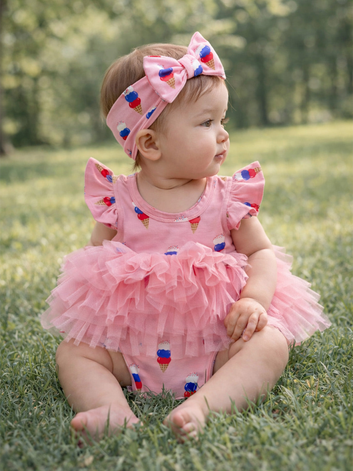 Baby girl in a pink ice cream-patterned dress and headband, sitting on grass, looking to the side.