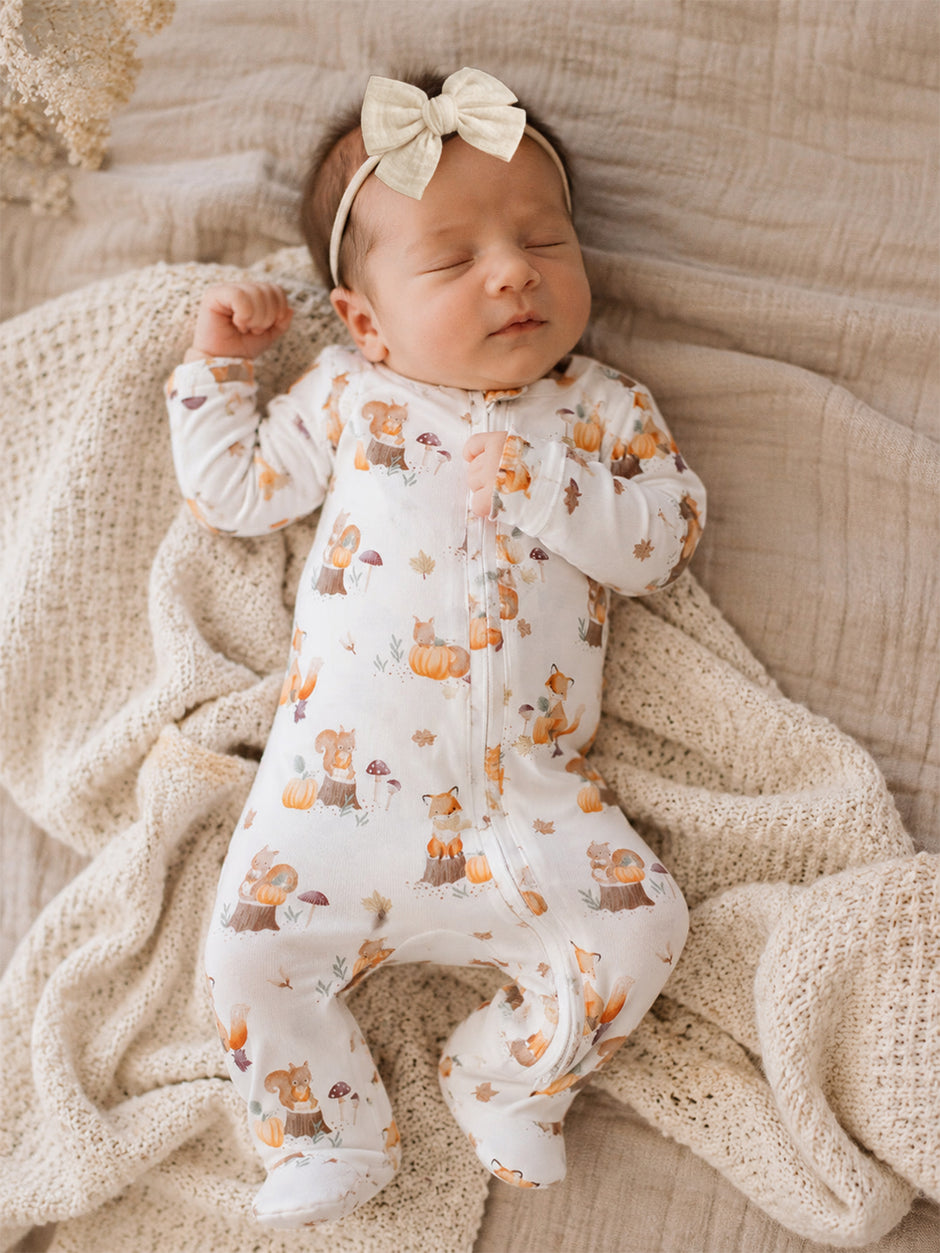 Baby wearing a pumpkin-patterned onesie and bow, peacefully sleeping on a textured blanket.