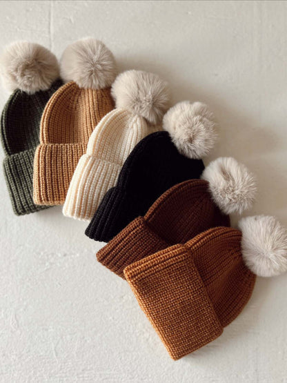 A collection of five cozy knit beanies in green, tan, cream, black, and brown, each featuring a fluffy pom-pom.