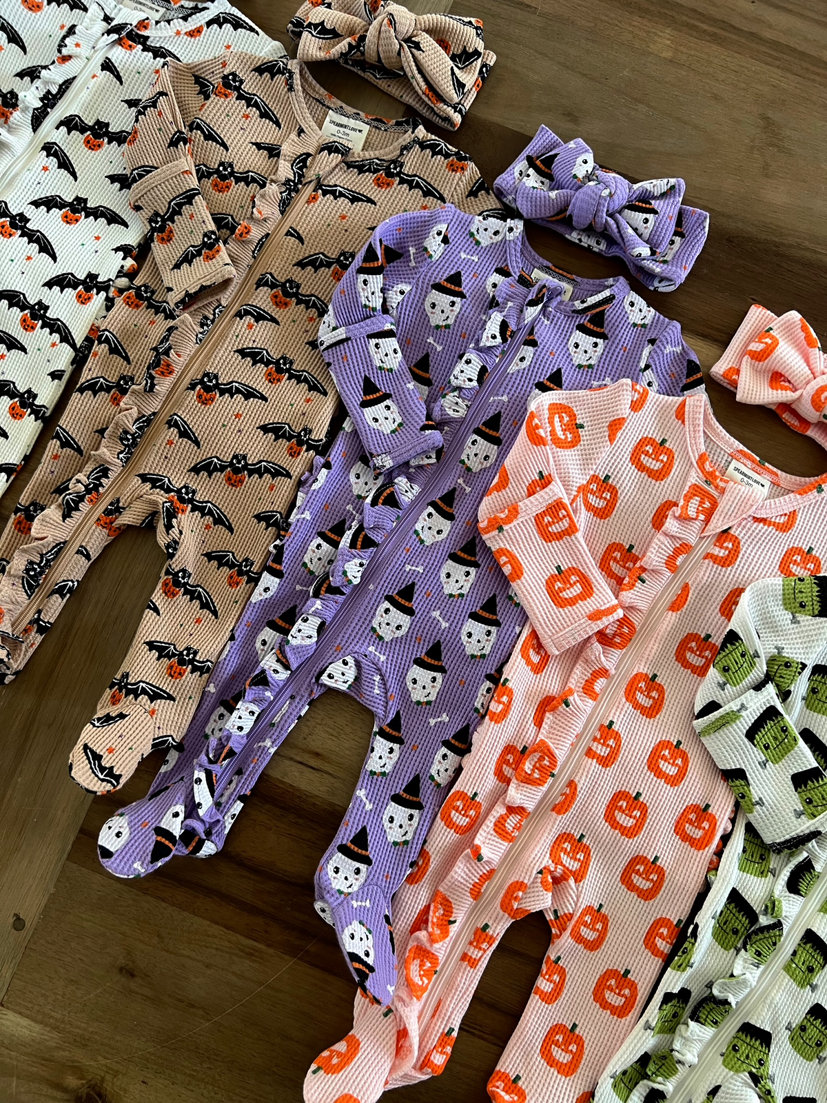 Colorful Halloween-themed baby onesies with matching headbands on a wooden surface.