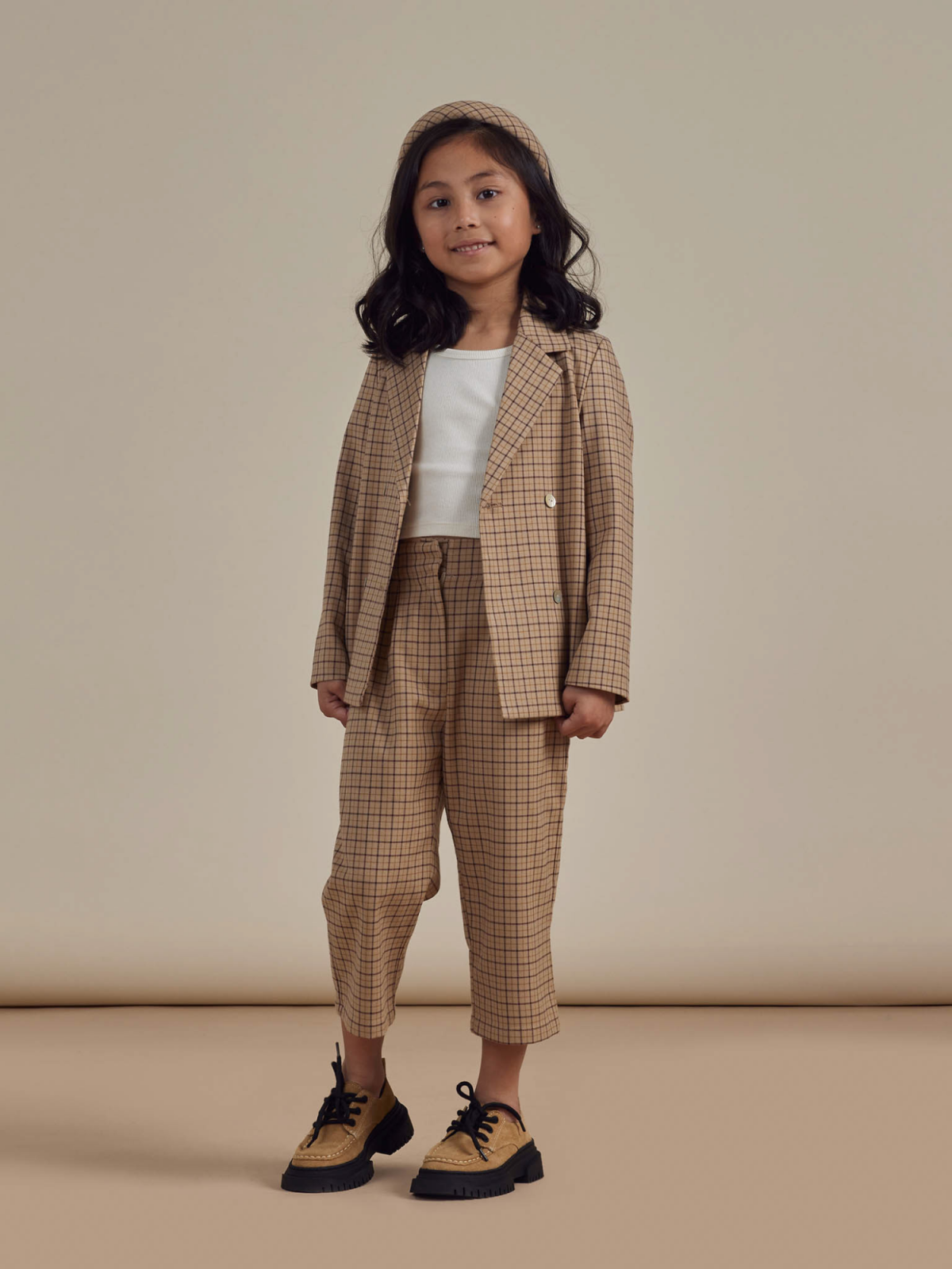 Girl in stylish plaid suit with a headband, standing against a neutral background, smiling confidently.