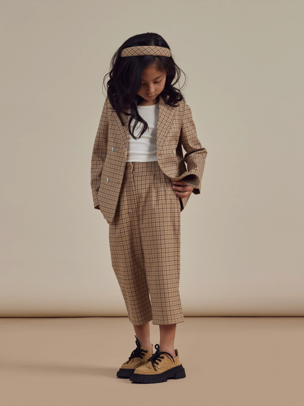 Child wearing a tan checkered suit, white top, and stylish shoes, looking down thoughtfully against a neutral background.