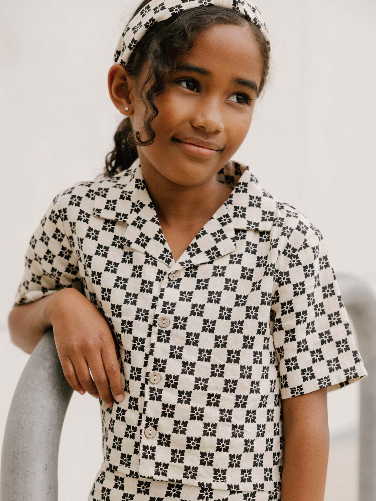 Young girl in patterned outfit, smiling and leaning on a railing, wearing a matching headband.