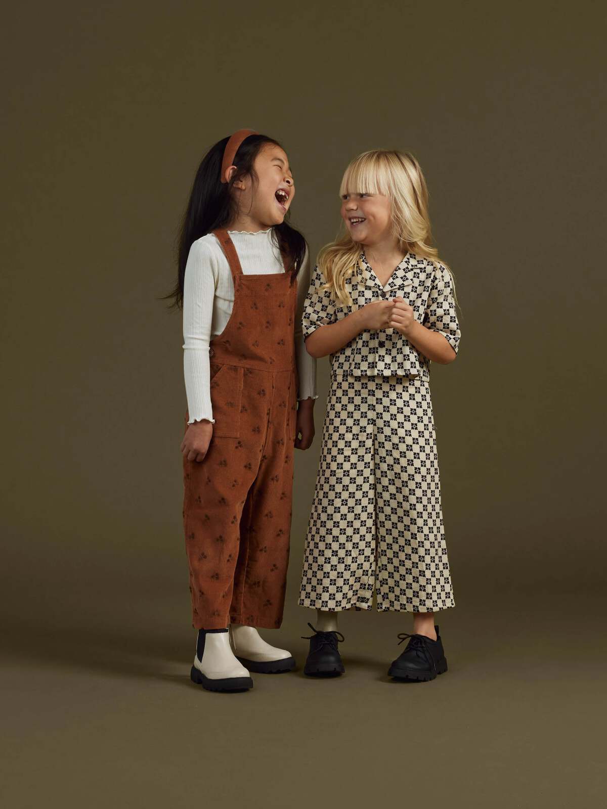 Two girls laughing while wearing stylish outfits against a muted background. One in an orange jumpsuit, the other in patterned attire.