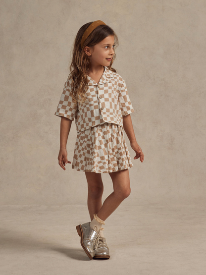 Child in a checked outfit with a matching headband, silver shoes, against a neutral background.