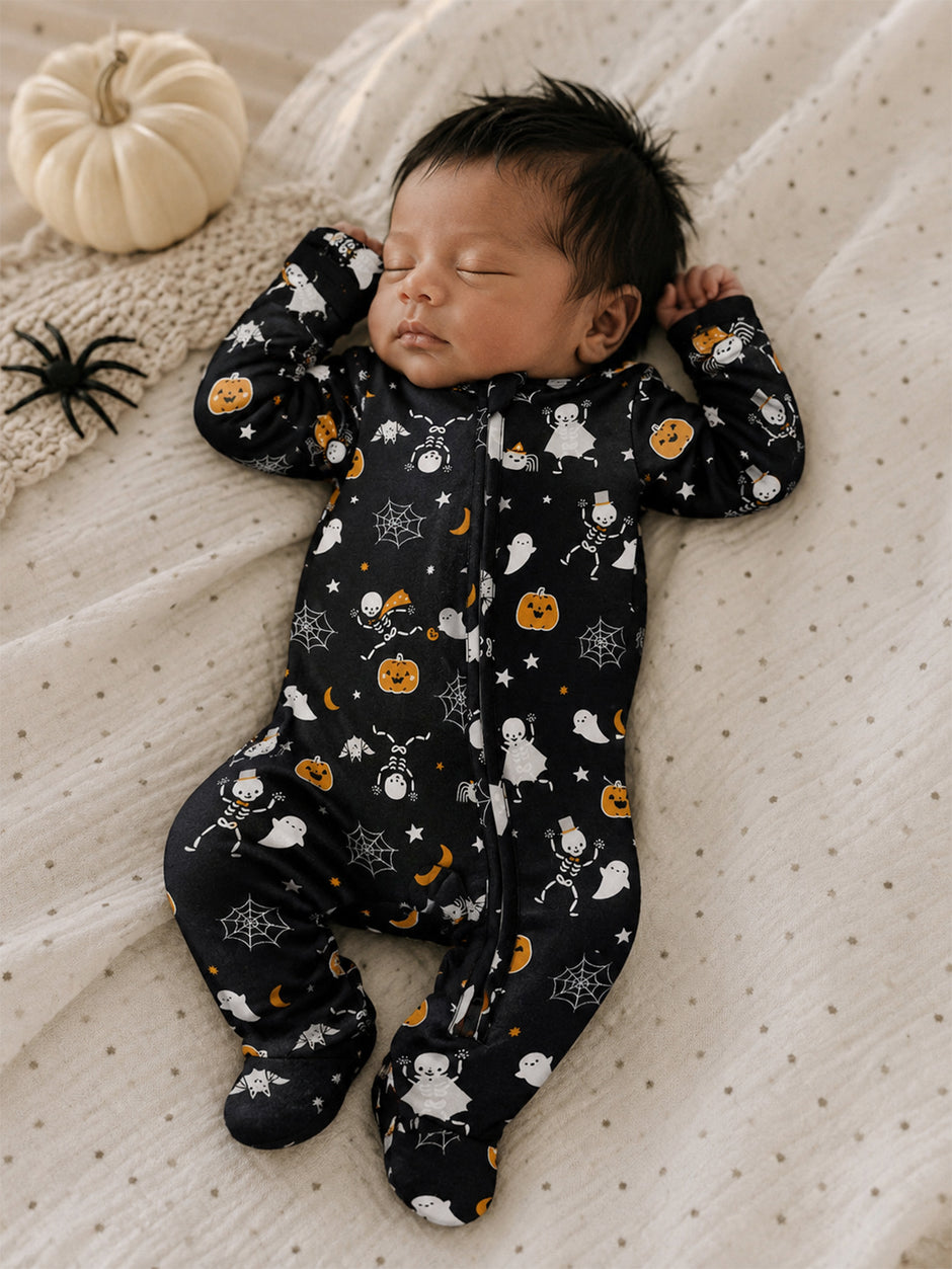 Baby sleeping peacefully in a Halloween-themed onesie with pumpkins, ghosts, and spiders. Cozy bedding in background.