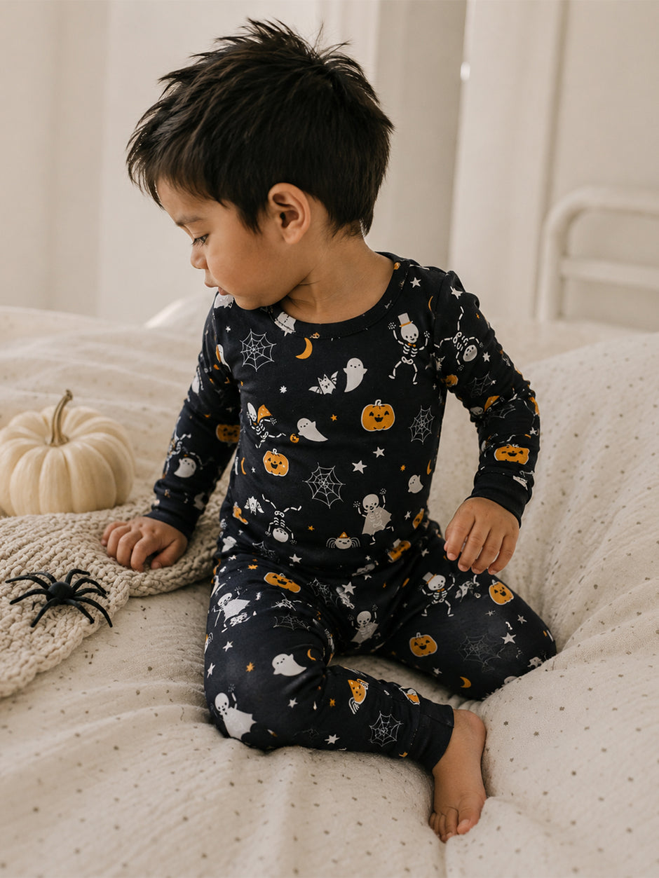 Child in Halloween-themed pajamas with ghosts and pumpkins, sitting on a bed next to a decorative pumpkin and spider.