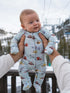 Baby in a colorful ski-themed onesie held up in snowy scenery with ski lift in the background.