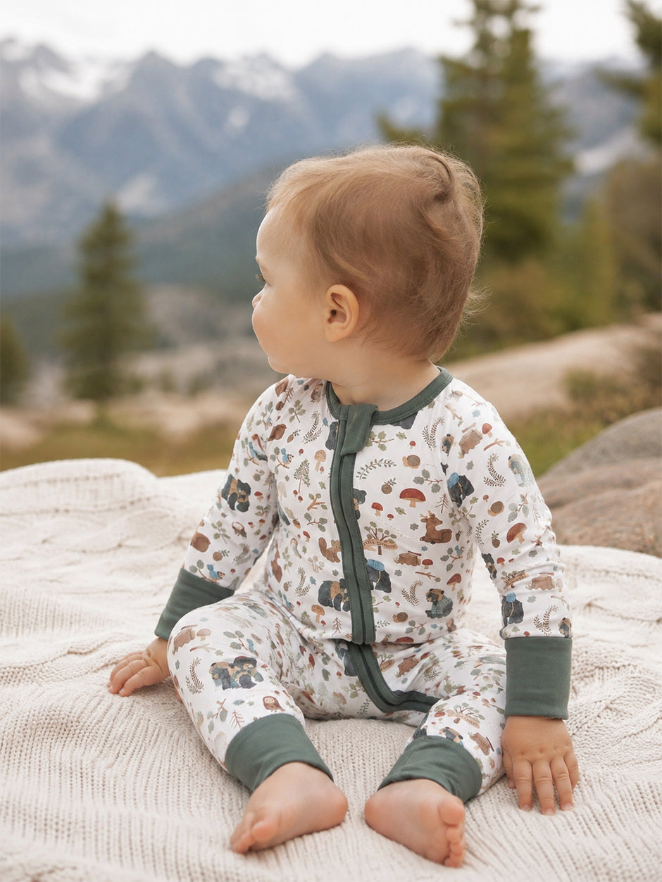 Toddler in nature-themed pajamas sitting on a blanket with mountains in the background.