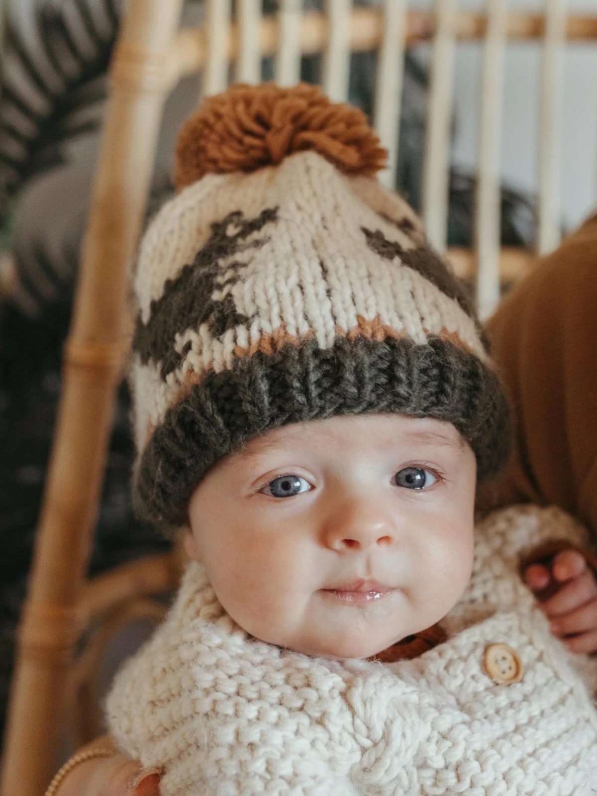 Baby in a knitted hat with a pom-pom, wearing a cream-colored sweater, sitting in a cozy environment.