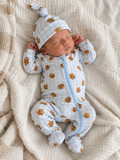 Newborn sleeping peacefully in a blue checkered onesie with bear prints and a matching hat on a cozy blanket.