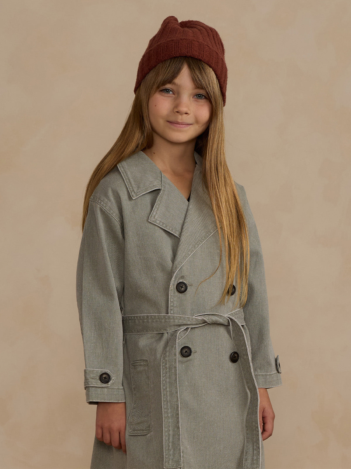 Girl wearing a gray coat and brown knitted hat, smiling against a neutral background.