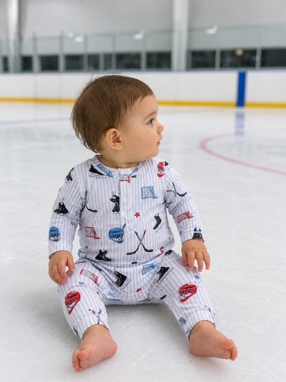 Baby in hockey-themed pajamas sitting on ice rink, looking to the side with a playful expression.