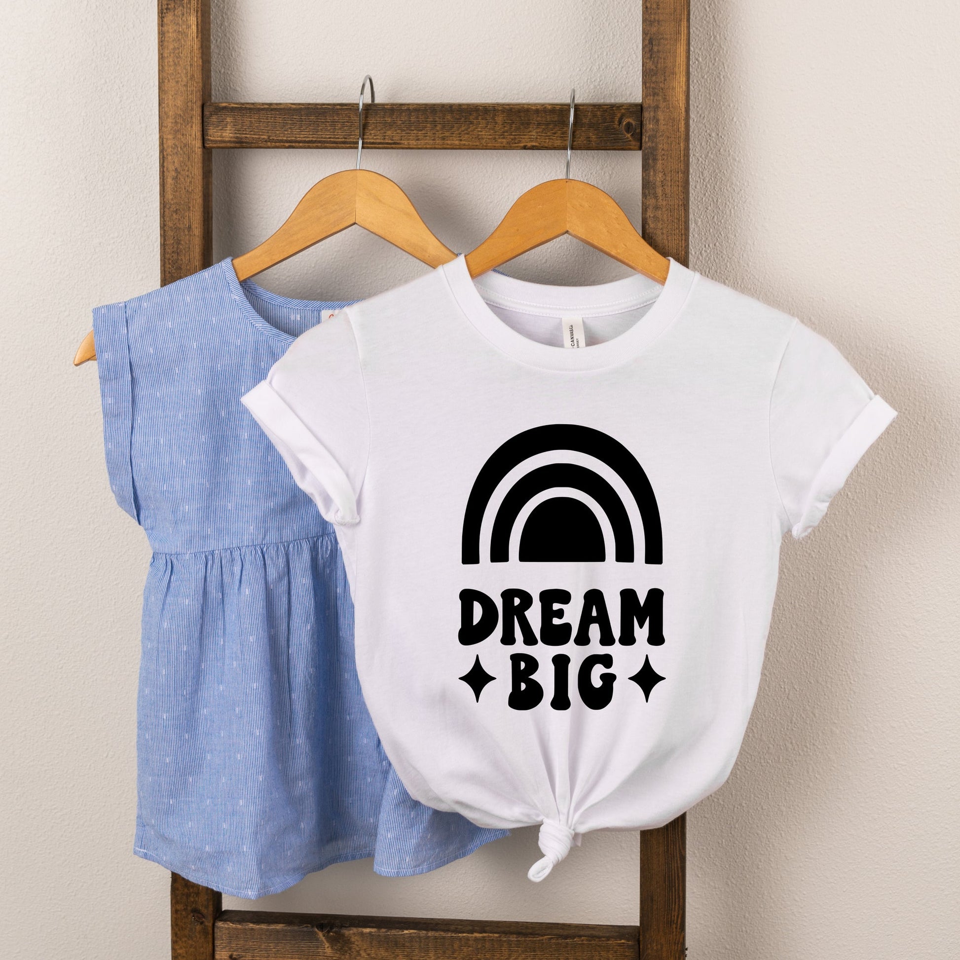 Two children's shirts hanging on a wooden rack: a blue top and a white shirt with a "Dream Big" rainbow design.