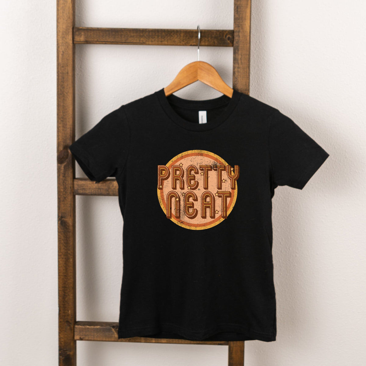 Black t-shirt hanging on a wooden ladder, featuring "Pretty Neat" graphic design in vibrant colors.