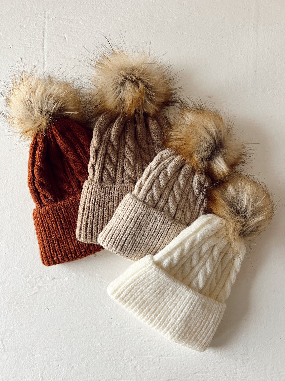 Three cozy knit beanies in rust, beige, and white with fluffy pom-poms on top against a textured white background.