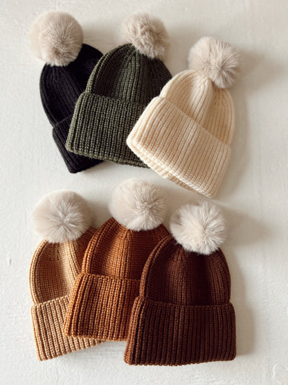A collection of ribbed knit beanies in black, green, cream, tan, and brown, each topped with a fluffy pom-pom.