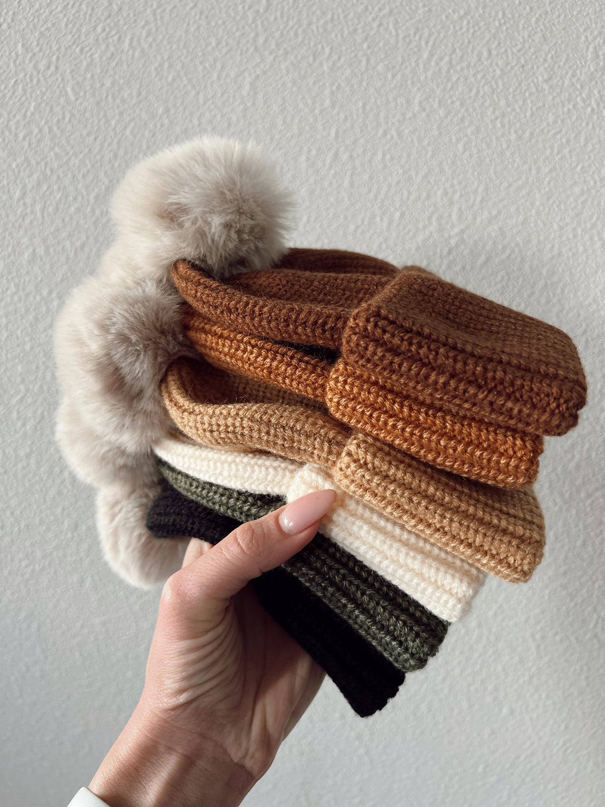 A hand holds a stack of knitted hats in earthy colors with fluffy pom-poms on top against a light background.