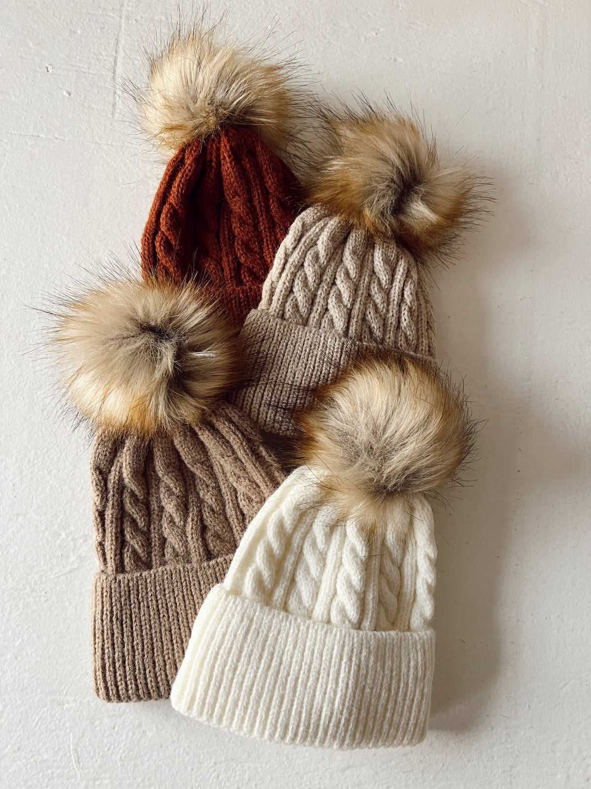 Four knitted beanies in various colors with fur pom-poms on top, arranged on a light background.