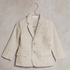 Light beige children's blazer with two buttons, hanging on a copper hook against a textured background.