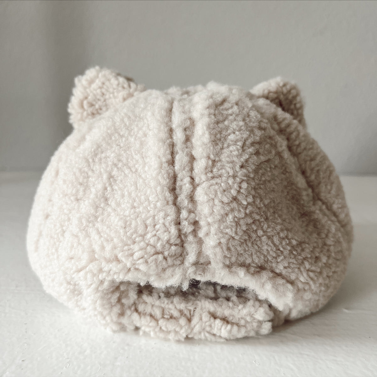 Beige fluffy hat with cat ears, displayed from the back on a light surface.