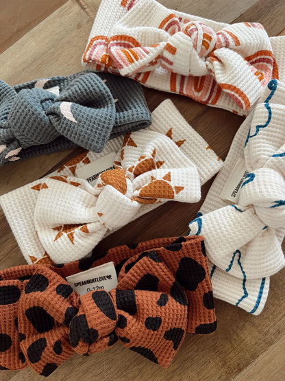 Six baby headbands arranged on a wooden surface, featuring various colors and patterns, including floral and animal prints.