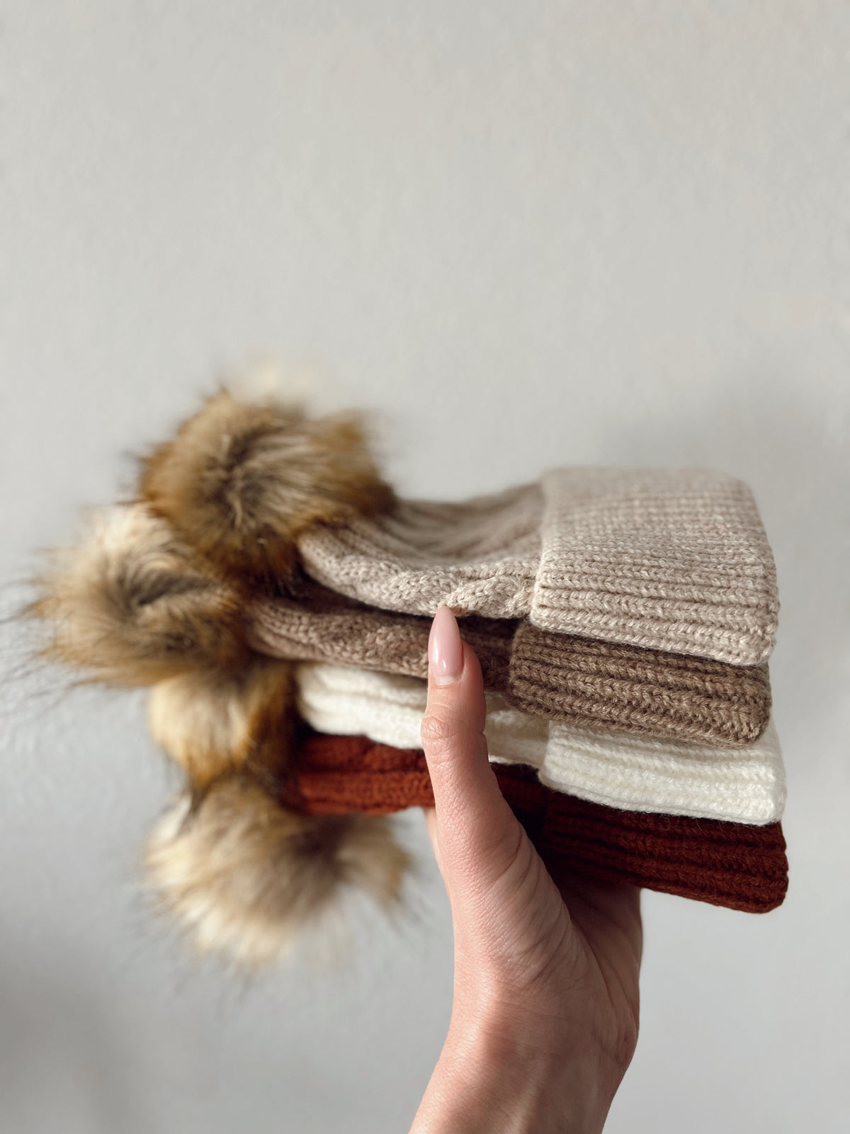 Hand holding a stack of cozy knit hats in neutral colors with fur pom-poms on top.